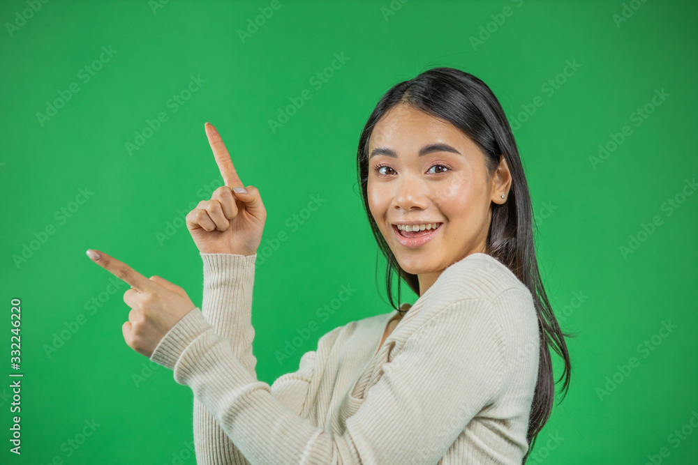 Portrait of beautiful 20s Asian woman wearing adorable dress expressing happiness pointing fingers sideways at copyspace isolated on green background Portrait of beautiful 20s Asian woman wearing adorable dress expressing happiness pointing fingers sideways at copyspace isolated on green background