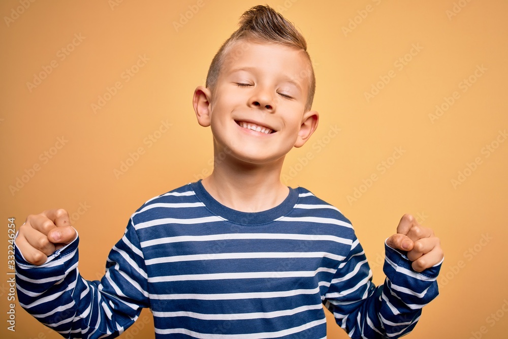 Young little caucasian kid with blue eyes wearing nautical striped shirt over yellow background very happy and excited doing winner gesture with arms raised, smiling and screaming for success Young little caucasian kid with blue eyes wearing nautical striped shirt over yellow background very happy and excited doing winner gesture with arms raised, smiling and screaming for success