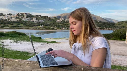 Close up of young blond girl freelance who remotely working on a laptop sitting on the bench with sea and mountain views
