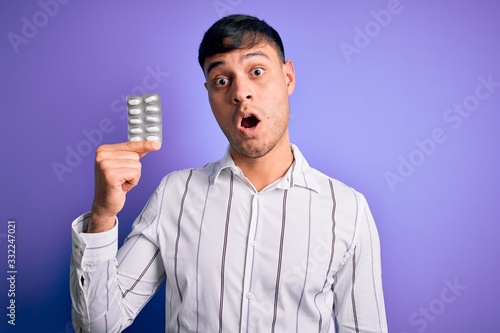 Young hispanic man holding pharmaceutical antibiotics pills over purple isolated background scared in shock with a surprise face, afraid and excited with fear expression