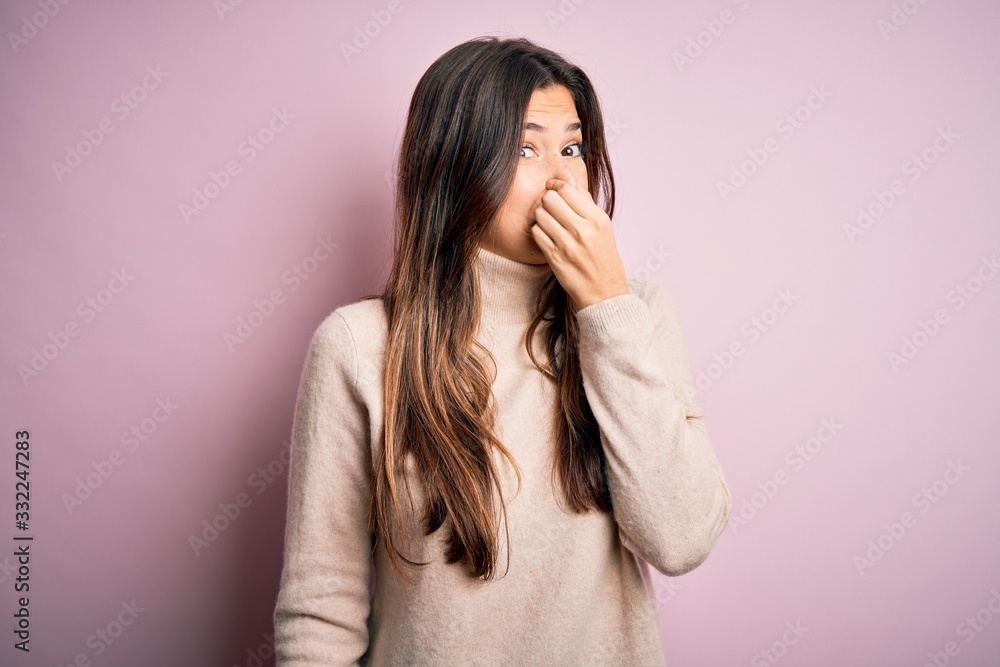 Young beautiful girl wearing casual turtleneck sweater standing over isolated pink background smelling something stinky and disgusting, intolerable smell, holding breath with fingers on nose. Bad Young beautiful girl wearing casual turtleneck sweater standing over isolated pink background smelling something stinky and disgusting, intolerable smell, holding breath with fingers on nose. Bad