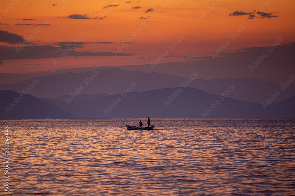 Romantic seascape. In the early morning or late evening, fishermen in a boat on a background of orange sky and purple mountains.