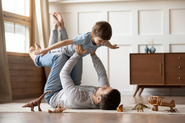 Happy young father lying on floor in living room hold fly with little preschooler son engaged in funny game together, loving dad relax playing with small boy child, enjoy family weekend at home