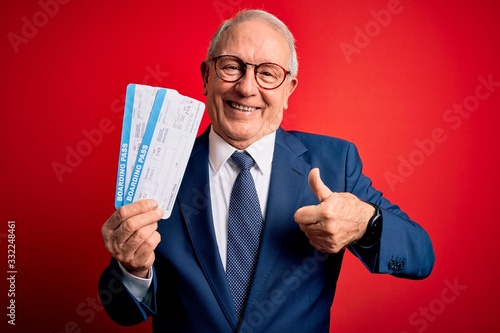 Senior grey haired business man holding airplane boarding pass over red background happy with big smile doing ok sign, thumb up with fingers, excellent sign