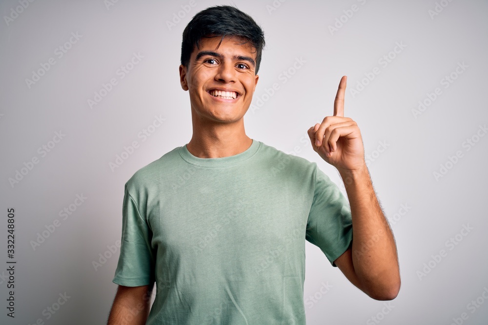 Fototapeta premium Young handsome man wearing casual t-shirt standing over isolated white background pointing finger up with successful idea. Exited and happy. Number one.