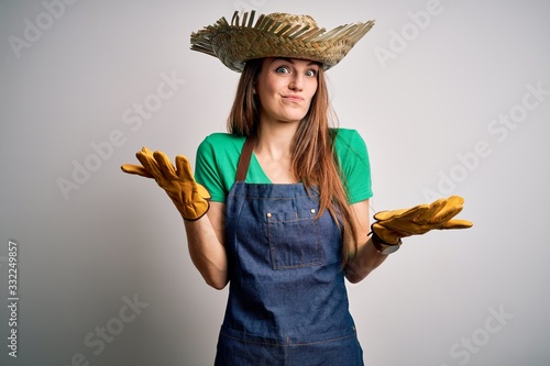 Young beautiful redhead farmer woman wearing apron and hat over white background clueless and confused expression with arms and hands raised. Doubt concept. photo