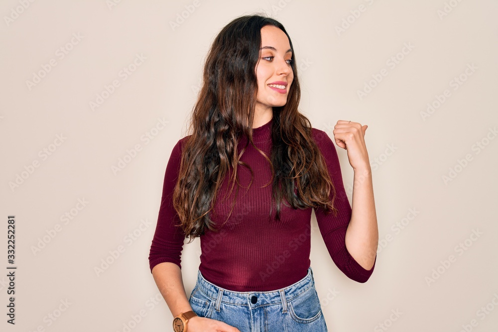 Young beautiful woman wearing casual t-shirt standing over isolated white background smiling with happy face looking and pointing to the side with thumb up. Young beautiful woman wearing casual t-shirt standing over isolated white background smiling with happy face looking and pointing to the side with thumb up.