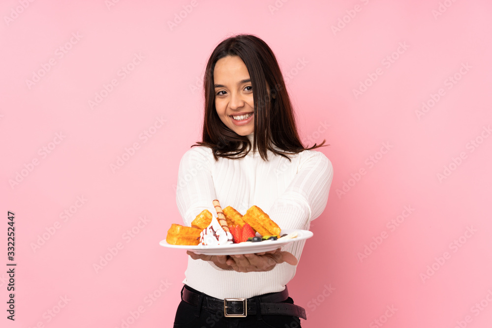 Young brunette woman holding waffles over isolated pink background holding copyspace imaginary on the palm to insert an ad Young brunette woman holding waffles over isolated pink background holding copyspace imaginary on the palm to insert an ad