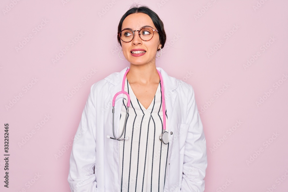 Beautiful doctor woman with blue eyes wearing coat and stethoscope over pink background looking away to side with smile on face, natural expression. Laughing confident. Beautiful doctor woman with blue eyes wearing coat and stethoscope over pink background looking away to side with smile on face, natural expression. Laughing confident.
