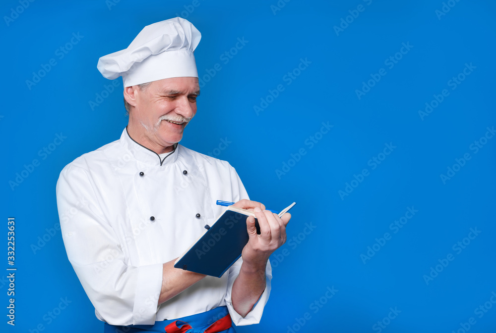 Photo of elderly master chef isolated on blue background, with his notepad, write new recipe Photo of elderly master chef isolated on blue background, with his notepad, write new recipe