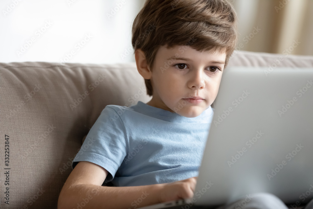 Concentrated smart little preschooler boy sit on sofa at home browsing wireless Internet on laptop, small serious child watch video use modern computer gadget, kids and technology, education concept Concentrated smart little preschooler boy sit on sofa at home browsing wireless Internet on laptop, small serious child watch video use modern computer gadget, kids and technology, education concept