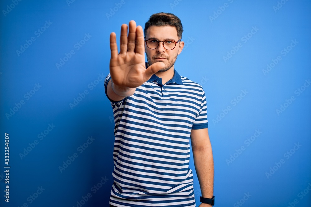 Naklejka premium Young man with blue eyes wearing glasses and casual striped t-shirt over blue background doing stop sing with palm of the hand. Warning expression with negative and serious gesture on the face.