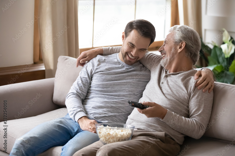 Smiling elderly father sit relax on couch with grown-up son have fun watching TV at home together, happy mature dad rest on sofa in living room with adult man child enjoy family weekend time