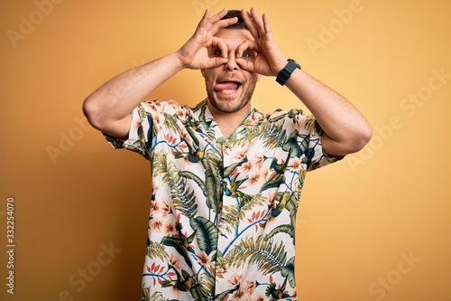 Young man with blue eyes on vacation wearing floral summer shirt over yellow background doing ok gesture like binoculars sticking tongue out  eyes looking through fingers. Crazy expression.