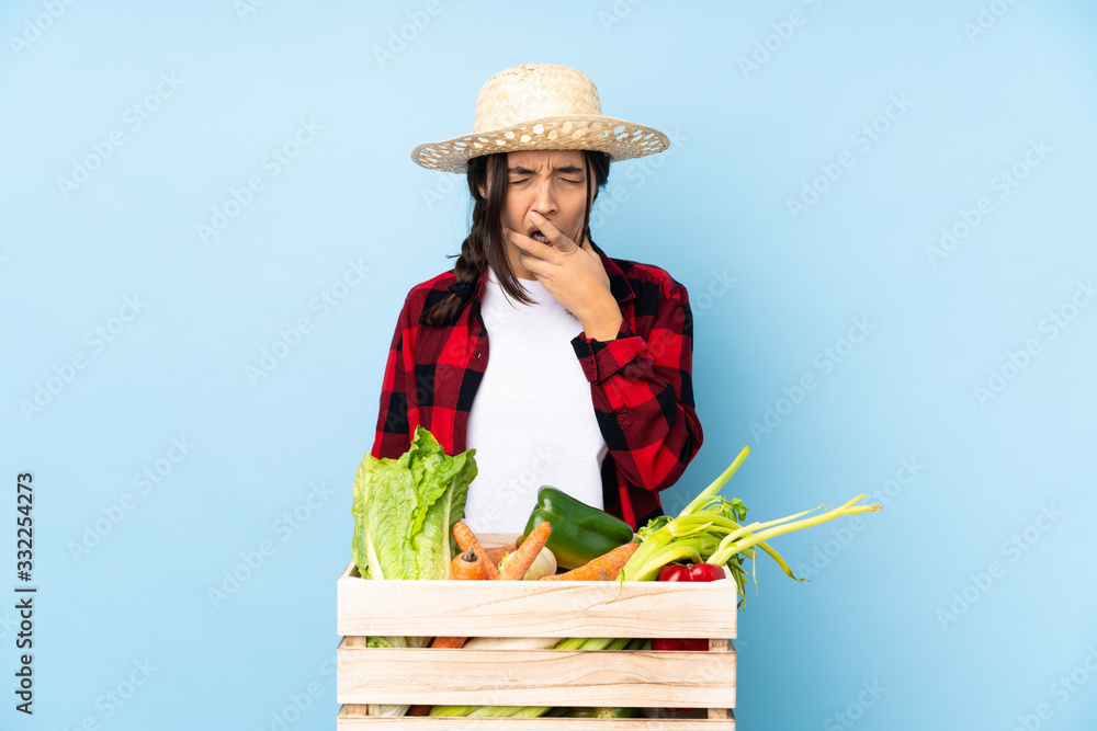 Young farmer Woman holding fresh vegetables in a wooden basket yawning and covering wide open mouth with hand