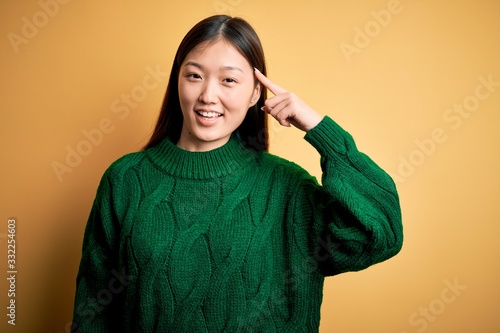 Young beautiful asian woman wearing green winter sweater over yellow isolated background Smiling pointing to head with one finger, great idea or thought, good memory © Krakenimages.com