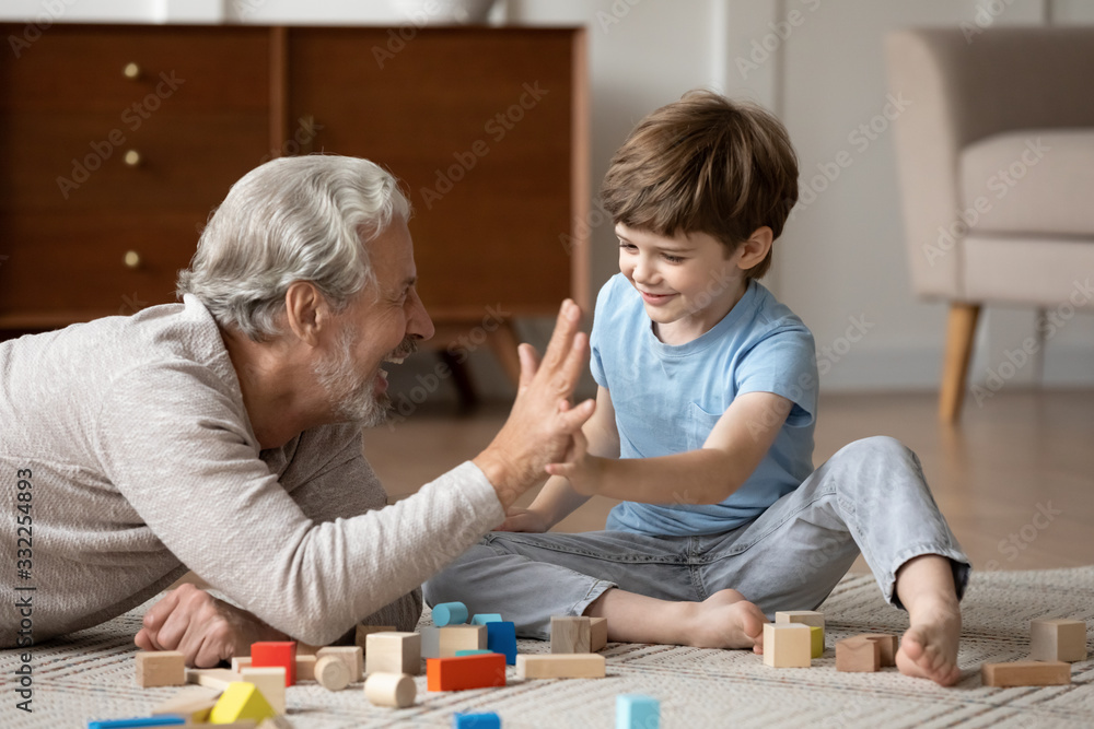 Overjoyed senior grandfather lying on floor in living room play with blocks with cute little grandson, happy mature grandparent relax engaged in game with building bricks with small grandchild at home - obrazy, fototapety, plakaty 