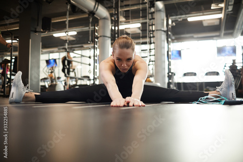 Front view portrait of blond young woman doing splits and stretching while enjoying workout in gym, copy space