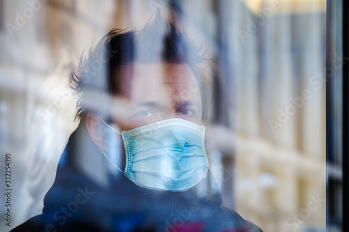Handsome European man stands behind glass in quarantine with a medical face mask on. Closeup of a 35-year-old male in a respirator to protect against infection with coronavirus (Covid-19). Defocused