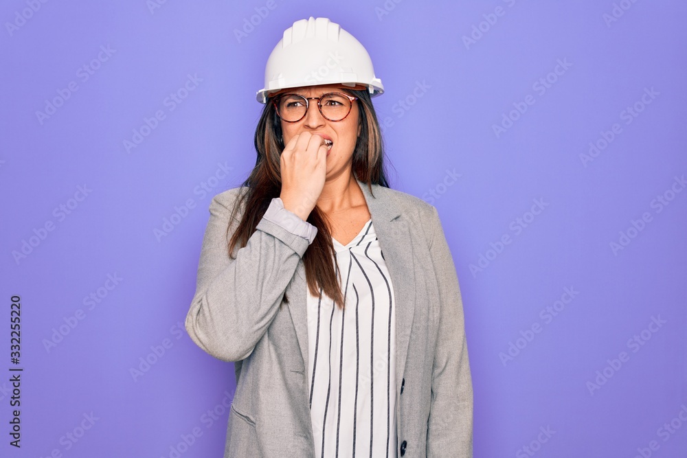 Professional woman engineer wearing industrial safety helmet over pruple background looking stressed and nervous with hands on mouth biting nails. Anxiety problem.