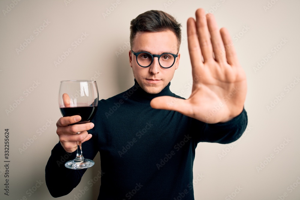 Young handsome caucasian man drinking an alcoholic glass of red wine over isolated background with open hand doing stop sign with serious and confident expression, defense gesture Young handsome caucasian man drinking an alcoholic glass of red wine over isolated background with open hand doing stop sign with serious and confident expression, defense gesture