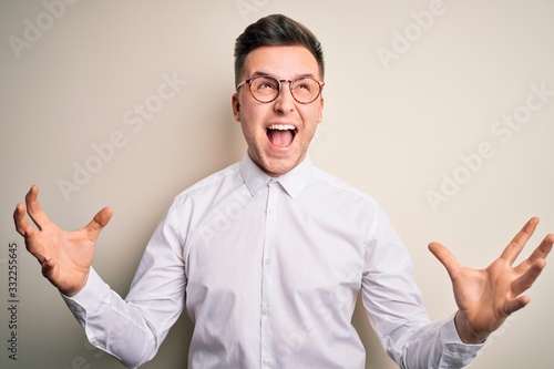 Young handsome business mas wearing glasses and elegant shirt over isolated background crazy and mad shouting and yelling with aggressive expression and arms raised. Frustration concept. © Krakenimages.com