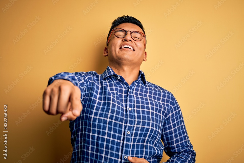 Young handsome latin man wearing casual shirt and glasses over yellow background laughing at you, pointing finger to the camera with hand over body, shame expression Young handsome latin man wearing casual shirt and glasses over yellow background laughing at you, pointing finger to the camera with hand over body, shame expression