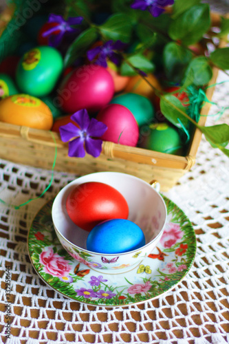 Easter colorful eggs, flowers and greens, white tea-set with eggs inside the cups with butterflies thin porcelain, on a white rounded knitted tablecloth. Country cozy style - happy holiday!