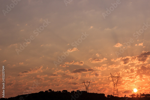 Mountain silhouette at sunrise. Sun rising between clouds on sky at morning. Pylons and trees on the mountain.