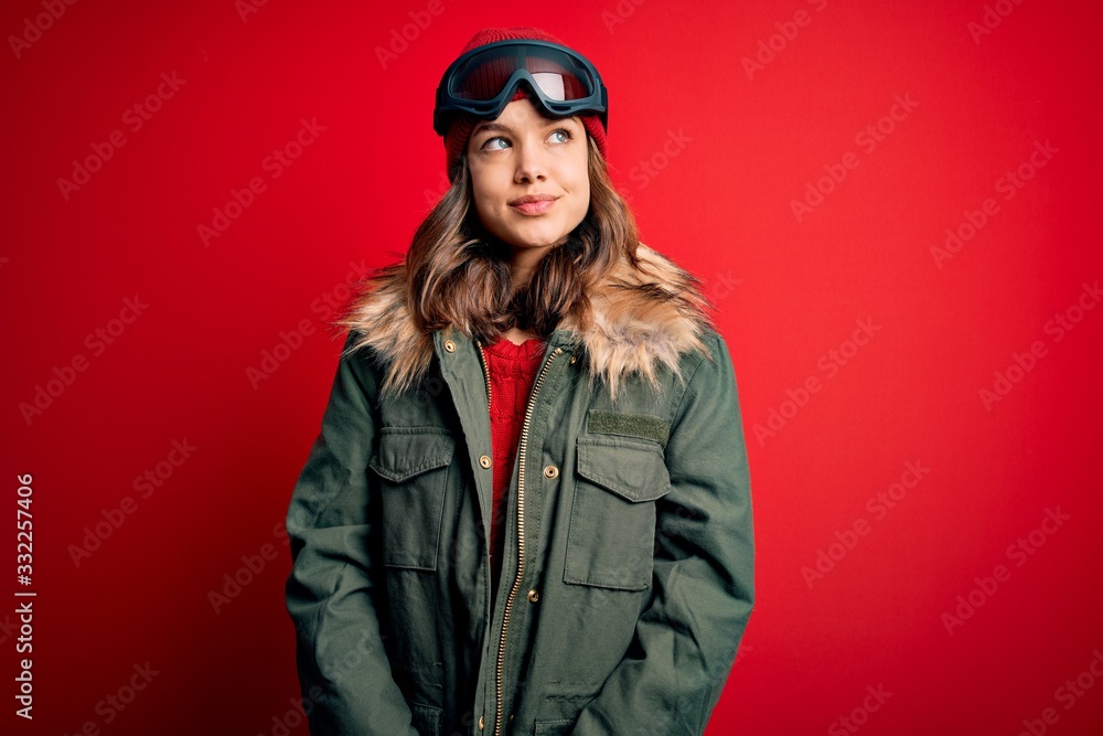 Young blonde girl wearing ski glasses and winter coat for ski weather over red background smiling looking to the side and staring away thinking.