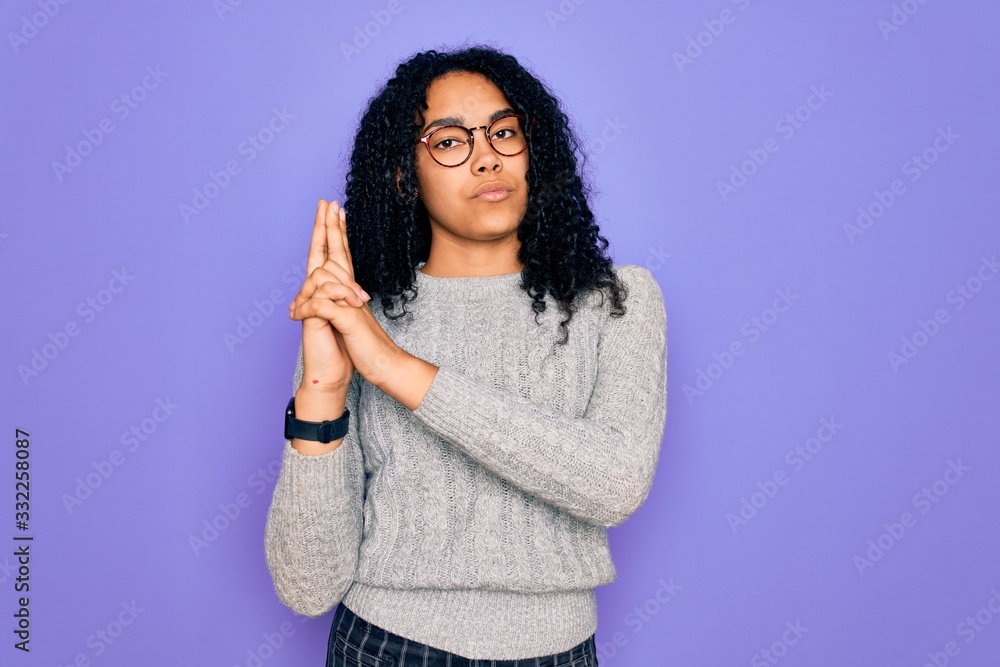 Young african american woman wearing casual sweater and glasses over purple background Holding symbolic gun with hand gesture, playing killing shooting weapons, angry face Young african american woman wearing casual sweater and glasses over purple background Holding symbolic gun with hand gesture, playing killing shooting weapons, angry face