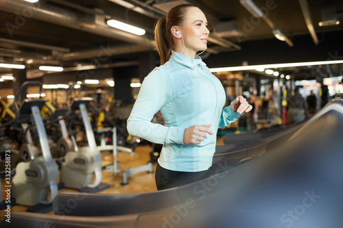 Side view portrait of cheerful young woman running on treadmill during cardio workout in modern gym, copy space