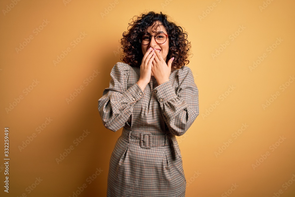 Beautiful arab business woman wearing dress and glasses standing over yellow background laughing and embarrassed giggle covering mouth with hands, gossip and scandal concept Beautiful arab business woman wearing dress and glasses standing over yellow background laughing and embarrassed giggle covering mouth with hands, gossip and scandal concept