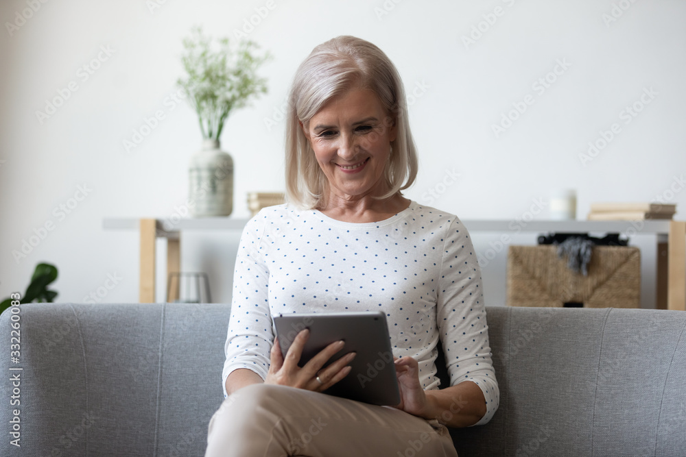 Smiling middle aged woman sitting on comfortable sofa with computer digital tablet. Happy older generation lady shopping online in internet store at home, using electronic device for communication. Smiling middle aged woman sitting on comfortable sofa with computer digital tablet. Happy older generation lady shopping online in internet store at home, using electronic device for communication.