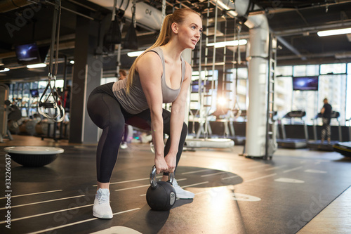 Full length portrait of muscular young woman picking up dumbbell during strength workout in sunlit gym, copy space