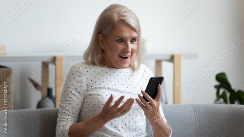 Excited mature woman holding mobile phone, received message with unexpected good news, sitting on couch at home. Happy middle aged grandma reading online lottery win email notification in smartphone.