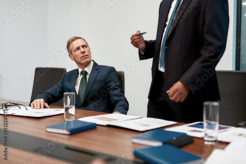 Interesting information. Portrait of serious mature businessman in formal wear listening to his colleague while sitting at the office table in the modern office