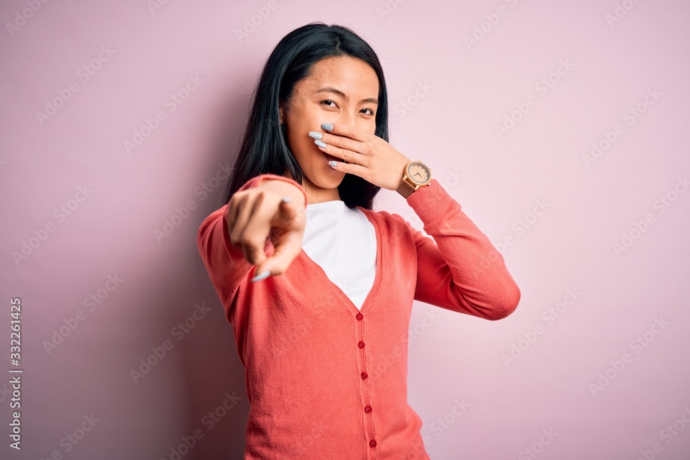 Young beautiful chinese woman wearing casual sweater over isolated pink background laughing at you, pointing finger to the camera with hand over mouth, shame expression Young beautiful chinese woman wearing casual sweater over isolated pink background laughing at you, pointing finger to the camera with hand over mouth, shame expression