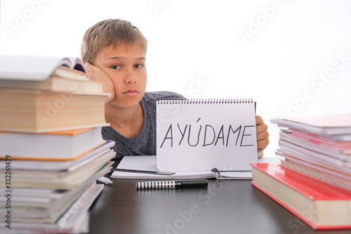 Tired frustrated boy sitting at the table with many books, exercises books. Spanish word Auydame - Help me- is written on open notebook. Learning difficulties, school, quarantine education concept photo