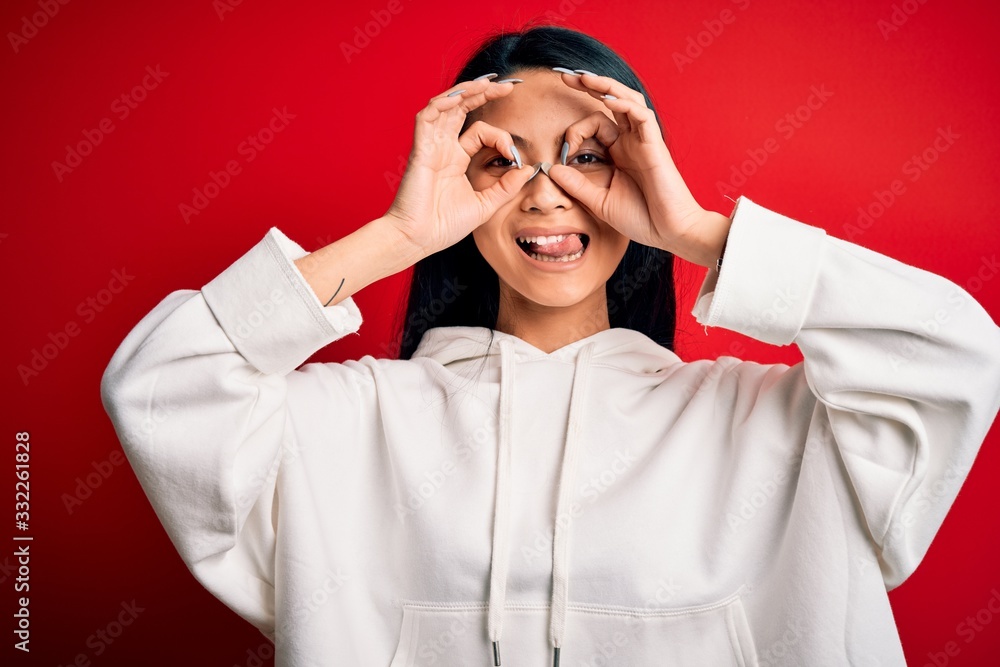 Young beautiful chinese sporty woman wearing sweatshirt over isolated red background doing ok gesture like binoculars sticking tongue out, eyes looking through fingers. Crazy expression. Young beautiful chinese sporty woman wearing sweatshirt over isolated red background doing ok gesture like binoculars sticking tongue out, eyes looking through fingers. Crazy expression.