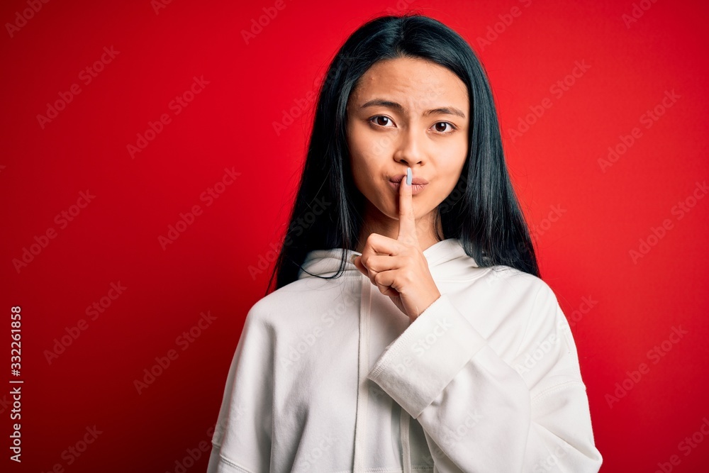 Young beautiful chinese sporty woman wearing sweatshirt over isolated red background asking to be quiet with finger on lips. Silence and secret concept. Young beautiful chinese sporty woman wearing sweatshirt over isolated red background asking to be quiet with finger on lips. Silence and secret concept.