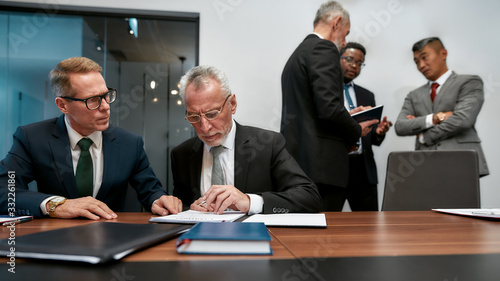 Signings documents. Confident mature businessman in eyeglasses reading a contract while having a meeting with his business partners in the modern office