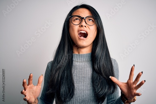 Young beautiful chinese woman wearing glasses and sweater over isolated white background crazy and mad shouting and yelling with aggressive expression and arms raised. Frustration concept.