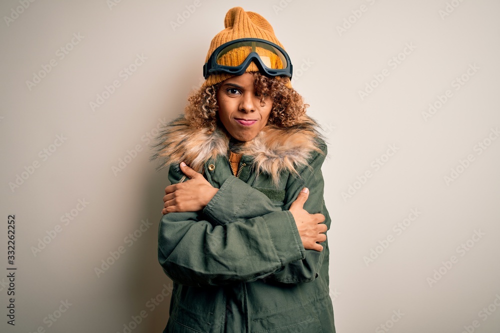 Young african american skier woman with curly hair wearing snow sportswear and ski goggles shaking and freezing for winter cold with sad and shock expression on face