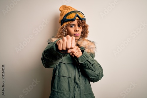 Young african american skier woman with curly hair wearing snow sportswear and ski goggles Punching fist to fight, aggressive and angry attack, threat and violence