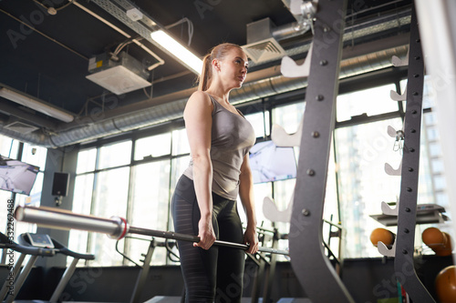 Low angle portrait of sportive young woman holding barbell during strength workout in modern gym, copy space