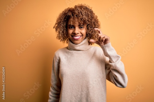 Young beautiful african american woman wearing turtleneck sweater over yellow background smiling and confident gesturing with hand doing small size sign with fingers looking and the camera. Measure © Krakenimages.com
