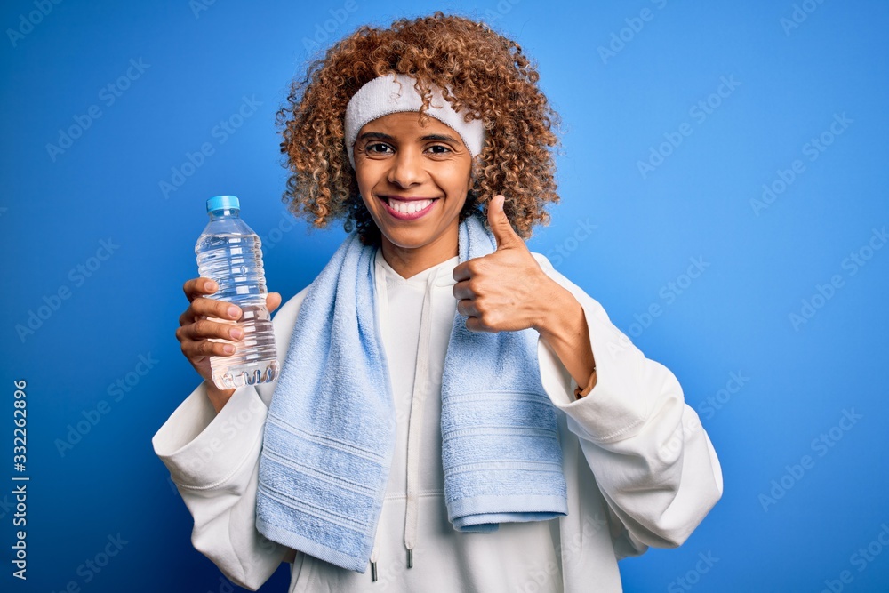 Young african american sportswoman doing sport wearing towel drinking botte of water happy with big smile doing ok sign, thumb up with fingers, excellent sign Young african american sportswoman doing sport wearing towel drinking botte of water happy with big smile doing ok sign, thumb up with fingers, excellent sign