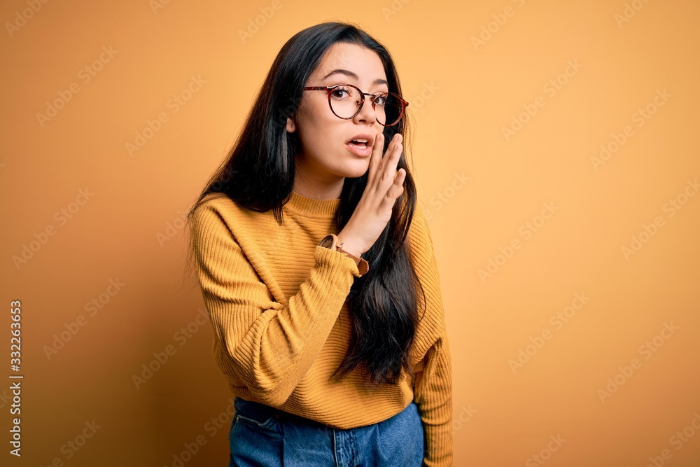 Young brunette woman wearing glasses and casual sweater over yellow isolated background hand on mouth telling secret rumor, whispering malicious talk conversation Young brunette woman wearing glasses and casual sweater over yellow isolated background hand on mouth telling secret rumor, whispering malicious talk conversation