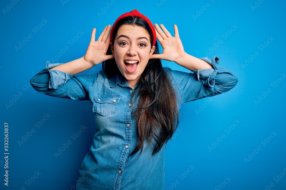 Young brunette woman wearing casual denim shirt over blue isolated background Smiling cheerful playing peek a boo with hands showing face. Surprised and exited Young brunette woman wearing casual denim shirt over blue isolated background Smiling cheerful playing peek a boo with hands showing face. Surprised and exited